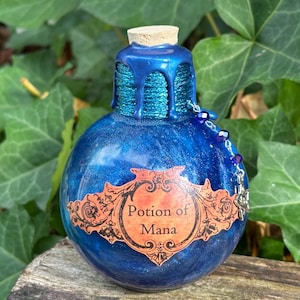 May include: A blue potion bottle with a cork stopper and a label that reads "Potion of Mana." The bottle has a textured, glittery finish and is adorned with a string of beads. The bottle is set against a backdrop of green leaves.