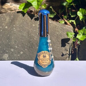 May include: A decorative glass bottle filled with blue liquid and sand. The bottle has a blue and brown top and a beaded chain. The label reads "AMPHITRITE DEPTH OF THE SEA".