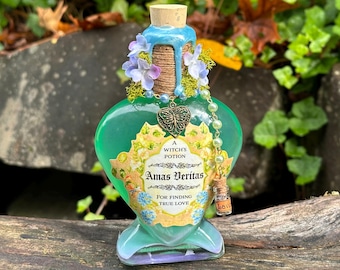 Könnte beinhalten: Eine herzförmige grüne Glasflasche mit Korkverschluss, umwickelt mit Schnur und blauem Wachs. Das Etikett trägt die Aufschrift A Witch's Potion Amas Veritas For Finding True Love und ist mit lila Blumen, Moos, einem Schmetterlingsanhänger und Perlen verziert.