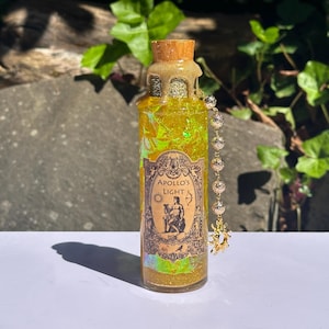May include: A decorative glass bottle filled with yellow liquid and glitter, featuring a cork stopper and a label that reads "Apollo's Light." A beaded charm hangs from the bottle. The bottle is approximately 6 inches tall.