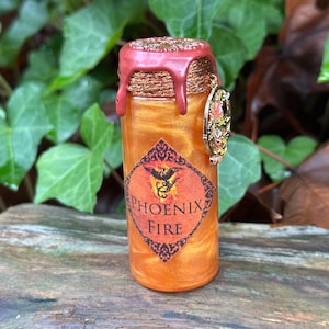 May include: A small, cylindrical glass bottle filled with a shimmering orange liquid. The bottle is sealed with a red wax top and a decorative gold charm. A label on the front reads "PHOENIX FIRE" with a phoenix illustration.
