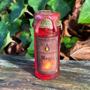 May include: A small, red glass bottle filled with glitter and a liquid. The bottle has a wax seal with gold accents and a label that reads "Sorcerer's Magic". The background features green leaves and brown foliage.