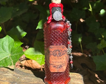 May include: A red glass bottle with a diamond-textured surface, sealed with a cork and red wax. A label reads Lilith's Defiance. A silver-toned charm featuring a circular emblem and a strand of red, black, and clear beads hangs from the bottle neck.