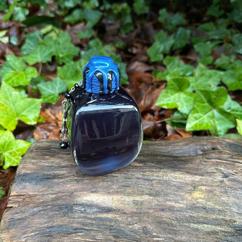 May include: A small, dark blue glass bottle with a black and blue stopper. The bottle has a silver chain with black beads attached to it.