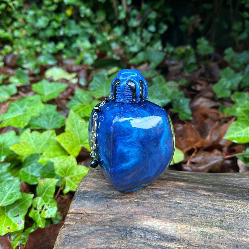 May include: A small, square, blue glass bottle with a black and silver stopper and a silver chain with black beads. The bottle has a glittery, iridescent finish.