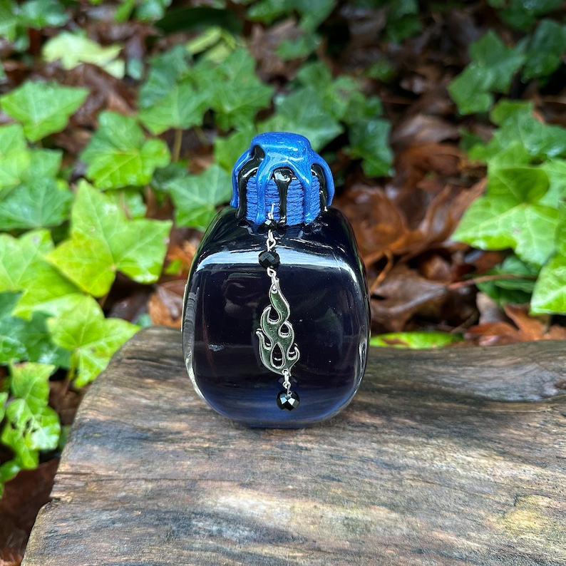 May include: A small, dark blue glass bottle with a black and blue dripping top. The bottle has a silver charm hanging from it, shaped like a flame.