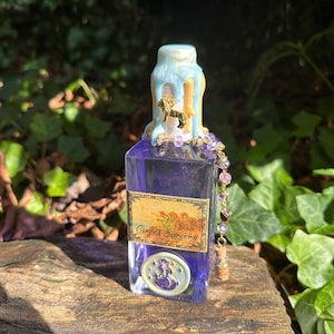 May include: A small glass bottle with a blue and white wax seal on the top. The bottle is filled with a purple liquid and has a label that says "Circe's Sorcery". A gold chain with purple beads hangs from the bottle.