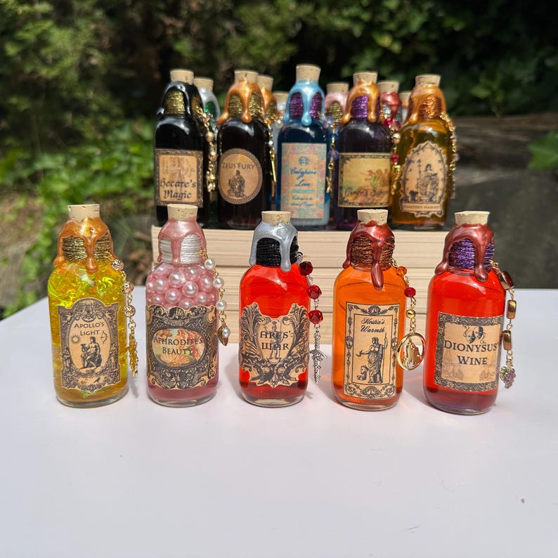 May include: A selection of decorative potion bottles with cork stoppers and detailed labels. The bottles are in various colours, including gold, red, and pink, with embellishments like beads and charms. Labels feature text such as "Apollo's Light" and "Aphrodite's Beauty."