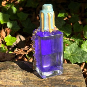 May include: A glass bottle with a purple liquid and a gold and blue wax-dipped stopper. The bottle has a gold chain with purple beads hanging from it.