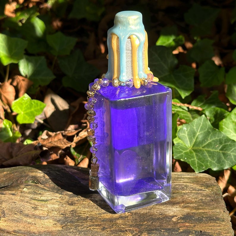 May include: A glass bottle with a purple liquid and a gold and blue wax-dipped stopper. The bottle has a gold chain with purple beads hanging from it.