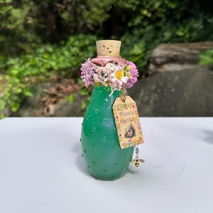 May include: A green glass bottle with a cork stopper, decorated with flowers and a tag reading "Flora's Garden." The bottle has a textured surface and a bee charm. The bottle is approximately 13 cm tall.