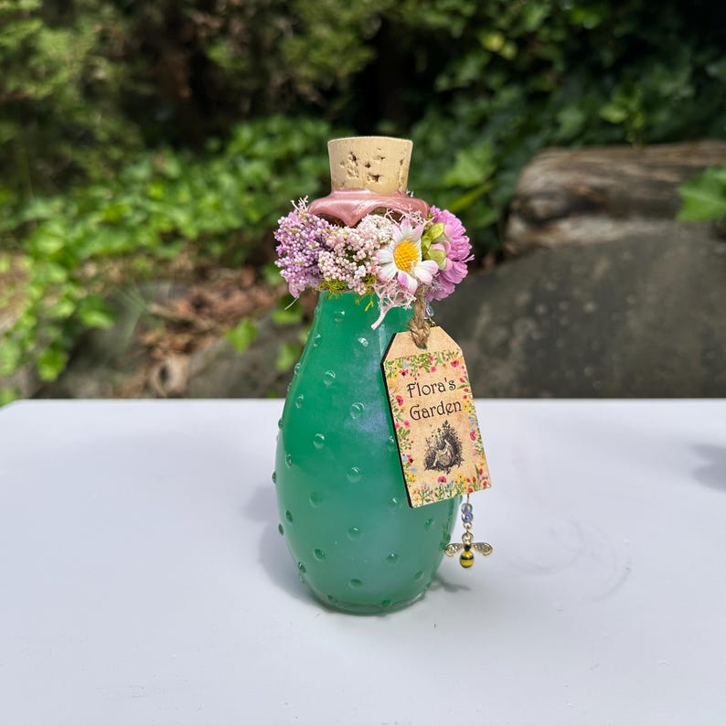 May include: A green glass bottle with a cork stopper, decorated with flowers and a tag reading "Flora's Garden." The bottle has a textured surface and a bee charm. The bottle is approximately 13 cm tall.