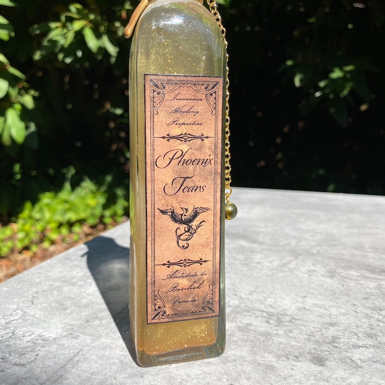 K&ouml;nnte beinhalten: Eine rechteckige Glasflasche, gef&uuml;llt mit einer schimmernden, goldenen Fl&uuml;ssigkeit. Die Flasche hat ein Etikett mit der Aufschrift "Phoenix Tears" und einer Phoenix-Illustration. Eine goldene Kette mit einer kleinen gr&uuml;nen Perle h&auml;ngt am Flaschenhals.