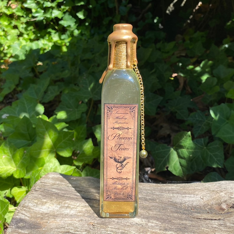 K&ouml;nnte beinhalten: Eine klare Glasflasche mit goldenem Wachssiegel und goldener Kette. Das Etikett tr&auml;gt die Aufschrift "Phoenix Tears" mit einer Phoenix-Illustration. Die Flasche enth&auml;lt eine goldene Fl&uuml;ssigkeit und steht auf Holz.