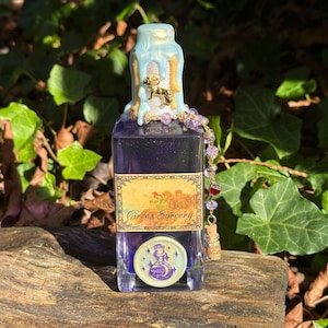 May include: A glass bottle with a blue and gold wax seal on the top. The bottle contains a purple liquid and has a label that reads "Circe's Sorcery". The bottle is decorated with a gold charm and a beaded chain.