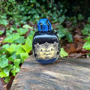 May include: A small, dark blue glass bottle with a blue and black stopper. The bottle has a gold label that reads "Hades' Kingdom".