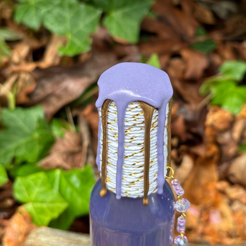K&ouml;nnte beinhalten: Eine violette Glasflasche mit einem gold-wei&szlig; gestreiften Design und einem violetten Wachssiegel oben. Die Flasche hat eine goldene Kette mit durchsichtigen Perlen, die daran h&auml;ngen.