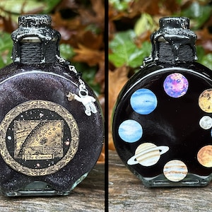 May include: A black glass bottle with a black wax seal and a silver astronaut charm. The bottle is decorated with a gold sticker that says "The Cosmos" and a black sticker with planets on it.