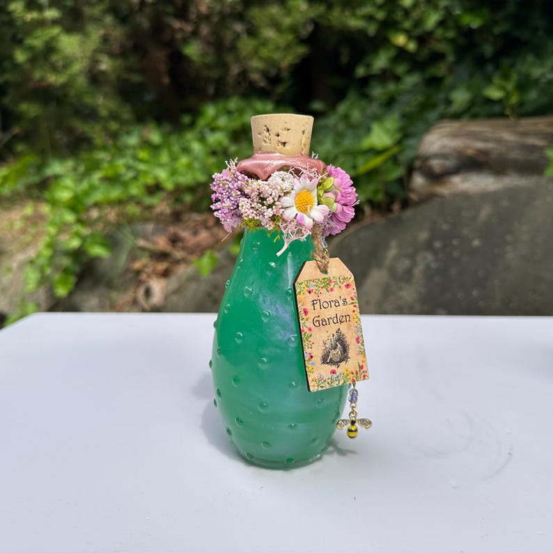 May include: A green glass bottle with a cork stopper, decorated with flowers and a tag reading "Flora's Garden." The bottle has a textured surface and a bee charm. The bottle is likely around 15 cm tall.