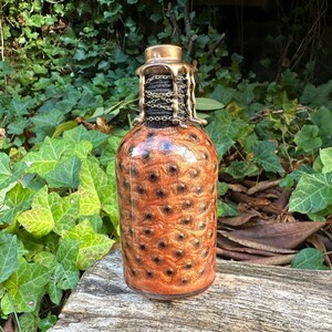 May include: A glass bottle with a gold-colored metal top and a brown and gold glitter design. The bottle has a chain wrapped around the neck.