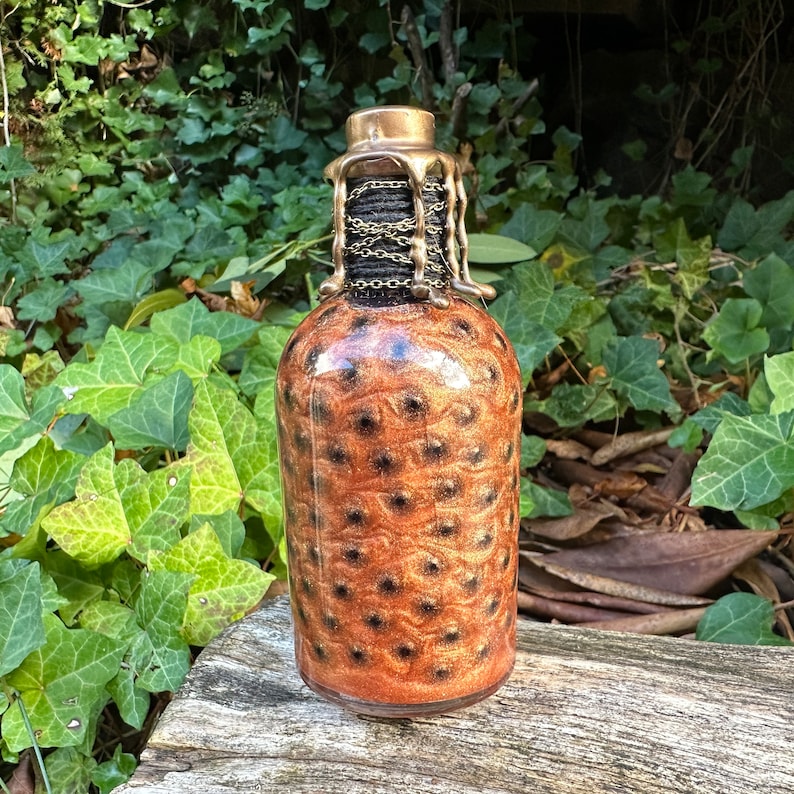 May include: A glass bottle with a gold-colored metal top and a brown and gold glitter design. The bottle has a chain wrapped around the neck.
