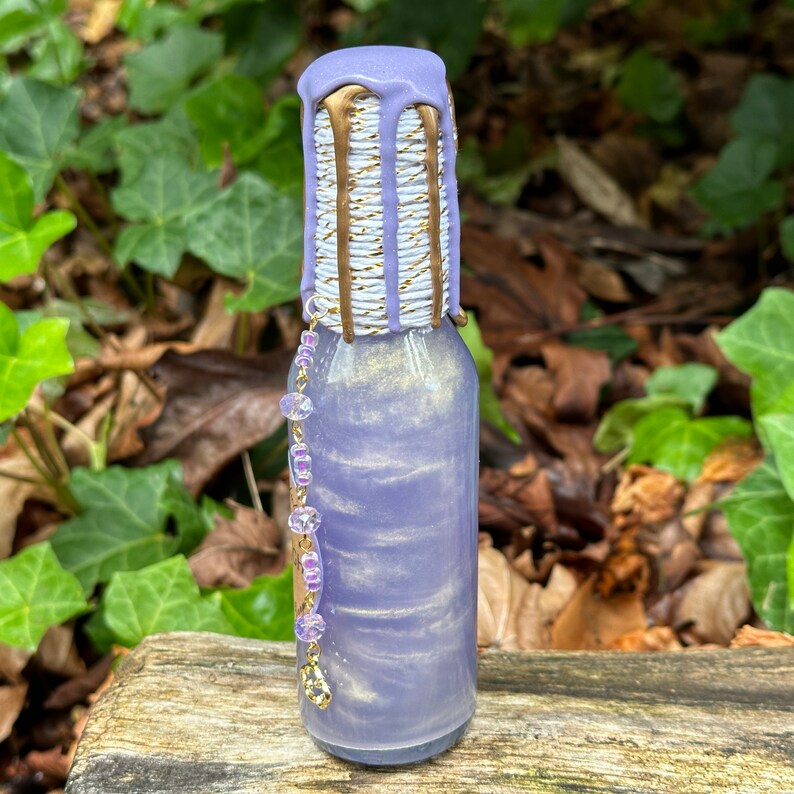 K&ouml;nnte beinhalten: Eine kleine Glasflasche mit einer lila und goldenen schillernden Fl&uuml;ssigkeit. Die Flasche hat einen wei&szlig;en und goldenen gestreiften Verschluss mit einem goldenen Tropfendesign. Eine lila und goldene Perlenkette h&auml;ngt an der Flasche.