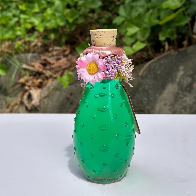 May include: A green glass bottle with a cork stopper, decorated with a pink flower and small purple flowers. The bottle has a textured surface with small, raised bubbles. The background is blurred green foliage.