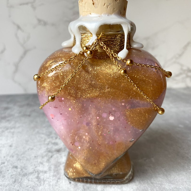 May include: A heart-shaped glass bottle with a gold glitter and pink liquid inside. The bottle has a gold chain with beads around the neck and a white wax seal on the top.