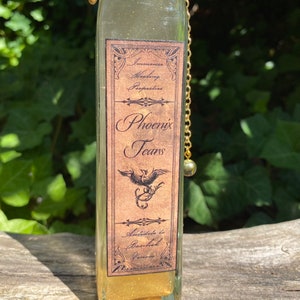 K&ouml;nnte beinhalten: Eine klare Glasflasche mit einer goldenen Kette und einem dekorativen Etikett. Das Etikett tr&auml;gt die Aufschrift "Phoenix Tears" mit einer Phoenix-Illustration. Die Flasche enth&auml;lt eine goldene Fl&uuml;ssigkeit, m&ouml;glicherweise ein Trank oder ein dekorativer Gegenstand.