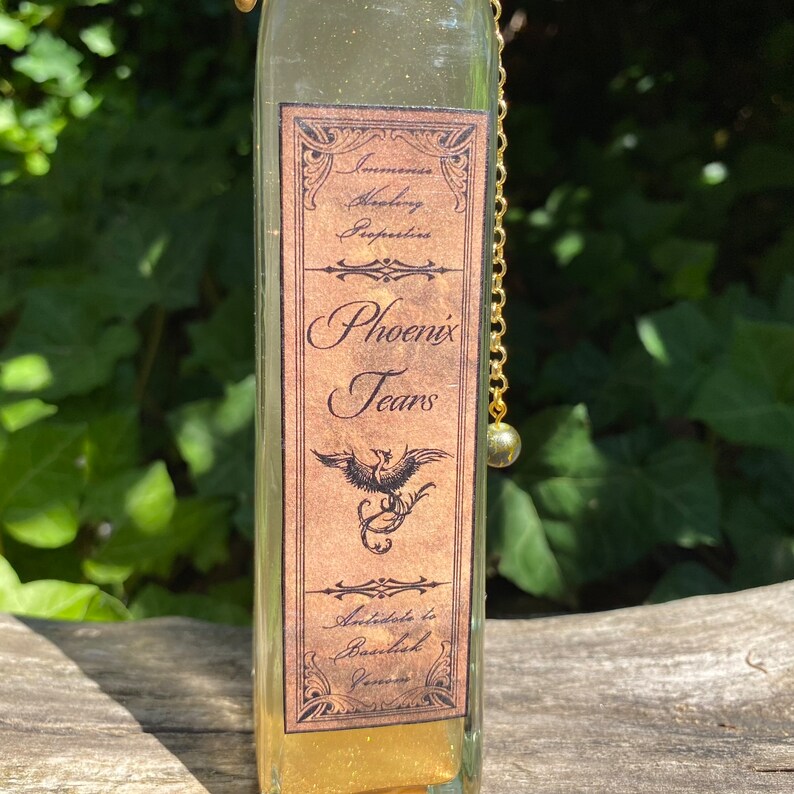 K&ouml;nnte beinhalten: Eine klare Glasflasche mit einer goldenen Kette und einem dekorativen Etikett. Das Etikett tr&auml;gt die Aufschrift "Phoenix Tears" mit einer Phoenix-Illustration. Die Flasche enth&auml;lt eine goldene Fl&uuml;ssigkeit, m&ouml;glicherweise ein Trank oder ein dekorativer Gegenstand.