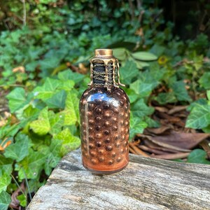 May include: A small glass bottle with a gold-colored metal cap and a chain around the neck. The bottle is filled with a glittery, copper-colored liquid.