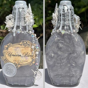 May include: A decorative glass bottle filled with a shimmering, silver liquid, labeled "Unicorn Blood." The bottle is adorned with silver accents, a beaded chain, and a unicorn charm. The top is decorated with silver drippings and white embellishments.