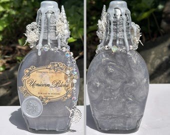 May include: A decorative glass bottle filled with a shimmering, silver liquid, labeled "Unicorn Blood." The bottle is adorned with silver accents, a beaded chain, and a unicorn charm. The top is decorated with silver drippings and white embellishments.