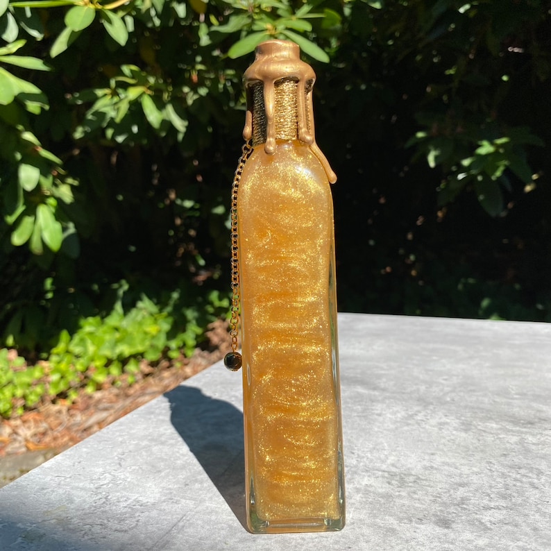 K&ouml;nnte beinhalten: Eine rechteckige Glasflasche gef&uuml;llt mit schimmernder goldener Fl&uuml;ssigkeit. Die Flasche hat einen goldfarbenen Verschluss mit strukturiertem Design und eine goldene Kette mit einem kleinen Anh&auml;nger. Die Flasche ist etwa 15 cm hoch.