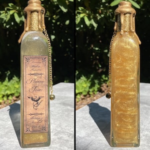 K&ouml;nnte beinhalten: Eine klare Glasflasche gef&uuml;llt mit schimmernder goldener Fl&uuml;ssigkeit, beschriftet mit "Phoenix Tears". Die Flasche hat ein goldfarbenes Wachssiegel und eine dekorative Kette mit einer Perle. Das Etikett ist braun mit verziertem Text und einer Ph&ouml;nix-Illustration.