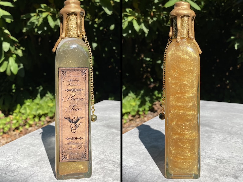 K&ouml;nnte beinhalten: Eine klare Glasflasche gef&uuml;llt mit schimmernder goldener Fl&uuml;ssigkeit, beschriftet mit "Phoenix Tears". Die Flasche hat ein goldfarbenes Wachssiegel und eine dekorative Kette mit einer Perle. Das Etikett ist braun mit verziertem Text und einer Ph&ouml;nix-Illustration.