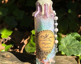 Peut inclure: Une bouteille en verre avec un sceau de cire rose et blanc, remplie de plumes irisées et d'une chaîne de perles. La bouteille porte une étiquette qui indique "Pegasus & Feathers".