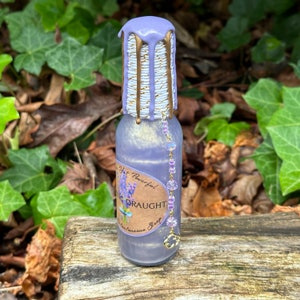K&ouml;nnte beinhalten: Eine kleine violette Glasflasche mit einem goldenen und wei&szlig;en Etikett, auf dem "Yet Powerful! Sleeping Draught For Instantaneous Sleep" steht. Die Flasche ist mit einem violetten Wachssiegel und einer Perlenkette versehen.