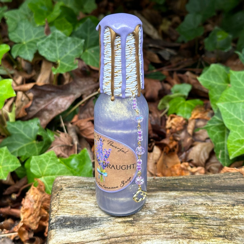 K&ouml;nnte beinhalten: Eine kleine violette Glasflasche mit einem goldenen und wei&szlig;en Etikett, auf dem "Yet Powerful! Sleeping Draught For Instantaneous Sleep" steht. Die Flasche ist mit einem violetten Wachssiegel und einer Perlenkette versehen.