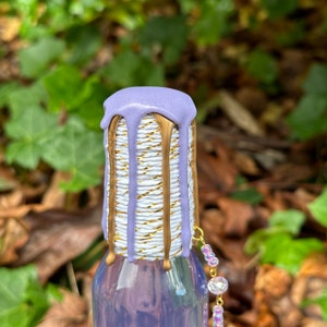 K&ouml;nnte beinhalten: Eine Glasflasche mit einem lila und goldenen Verschluss. Die Flasche ist mit einem wei&szlig;en und goldenen Seil verziert und hat einen lilafarbenen Tropfeffekt. Die Flasche hat einen klaren Glask&ouml;rper und eine goldene Kette mit durchsichtigen Perlen.