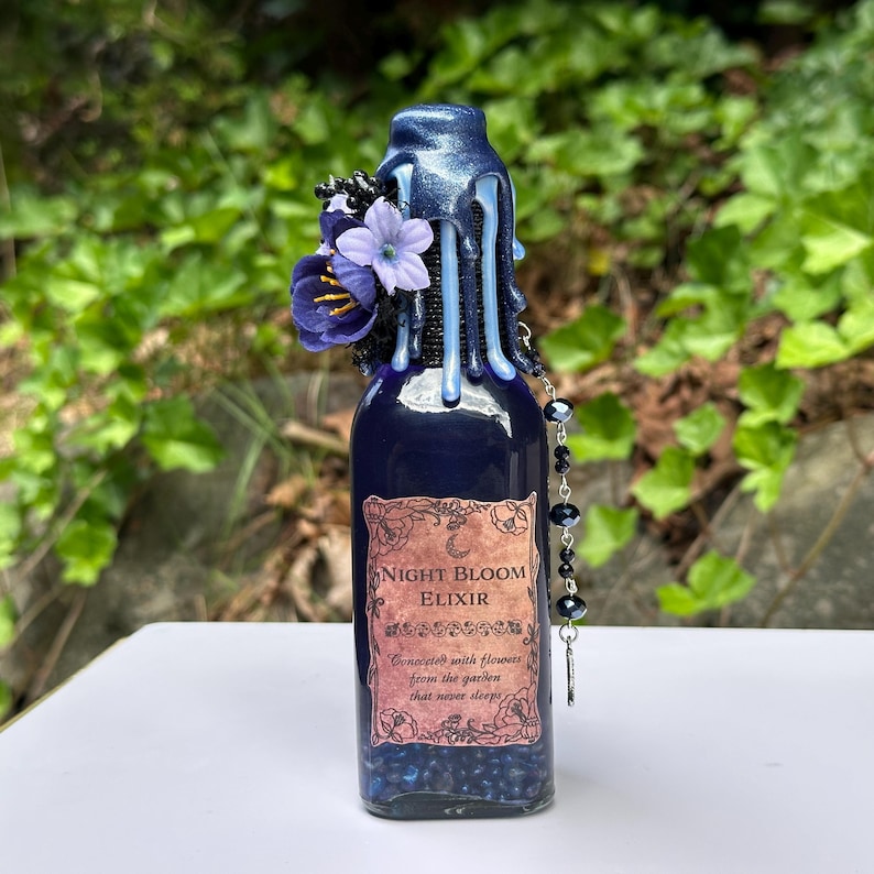 May include: A dark blue glass bottle with a decorative wax seal and floral accents. The bottle is labelled "Night Bloom Elixir" with a floral design. A beaded charm hangs from the bottle. The background is blurred green foliage.