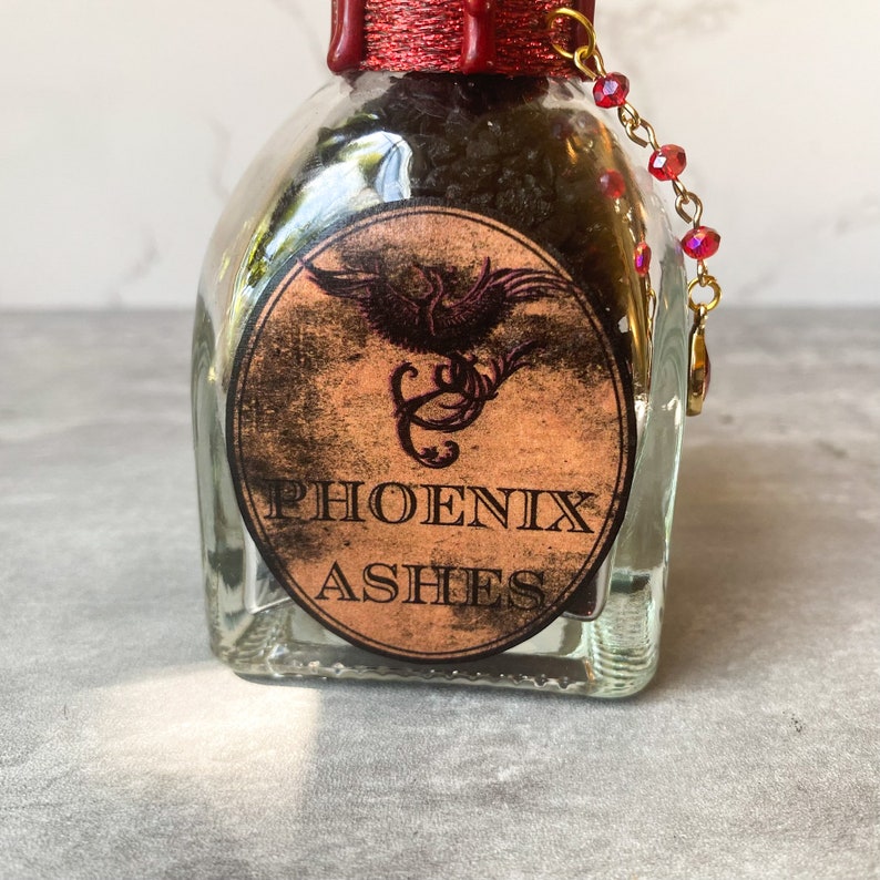 Puede incluir: Un frasco de vidrio con un sello de cera roja y una cadena dorada con cuentas rojas. El frasco contiene polvo negro y tiene una etiqueta que dice "PHOENIX ASHES" con un dise&ntilde;o de ave f&eacute;nix.