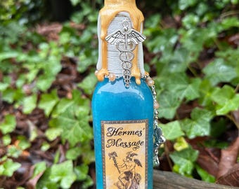 May include: A small blue glass bottle with a gold and white wax seal. The bottle has a silver charm with a snake and a label that reads "Hermes' Message".