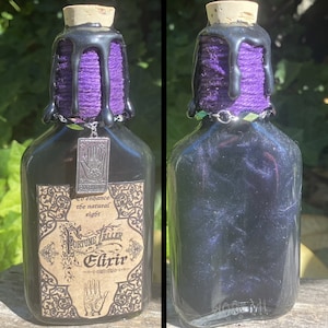 May include: A black glass bottle with a purple and black cork stopper. The bottle has a label that reads "Fortune Teller Elixir" and "To enhance the natural sight". The bottle is decorated with a silver charm with a hand design.