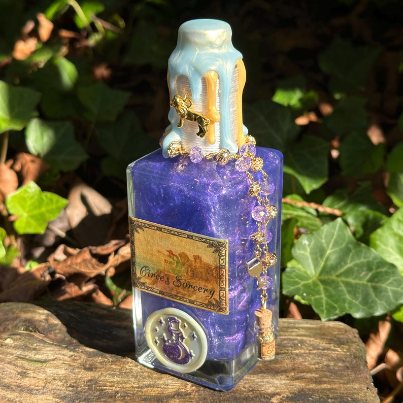 May include: A small glass bottle with a purple liquid and gold glitter inside. The bottle has a blue and gold wax seal on the top and a gold chain with purple beads hanging from it. The label on the bottle reads "Circe's Sorcery".