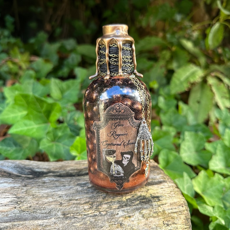 May include: A small glass bottle with a gold metal top and a black chain. The bottle is filled with a dark liquid and has a label that reads 'Gin Reaper's Captured Souls'. A silver skeleton hand charm hangs from the chain.