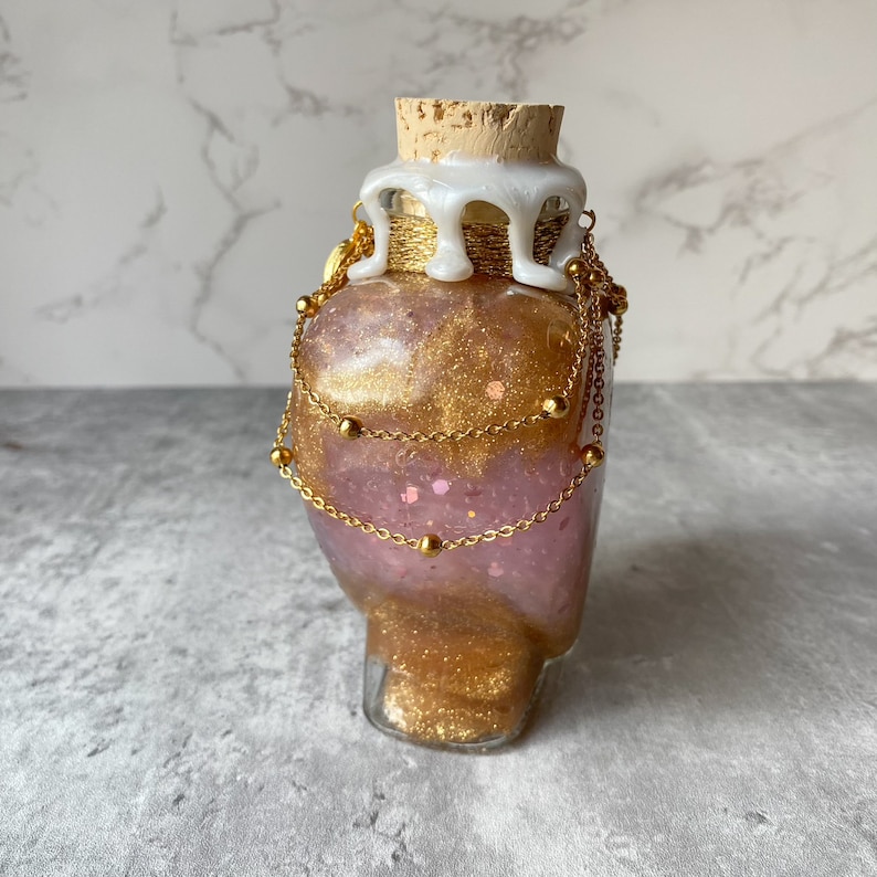 May include: A glass bottle with a cork stopper and a white wax seal. The bottle is filled with pink and gold glitter and has gold chains wrapped around it.