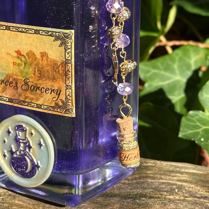 May include: A purple glass bottle with a gold label that reads "'s Sorcery". The bottle has a gold charm attached to it with purple and gold beads. The charm is a small glass bottle with a cork stopper and a label that reads "Herbs".