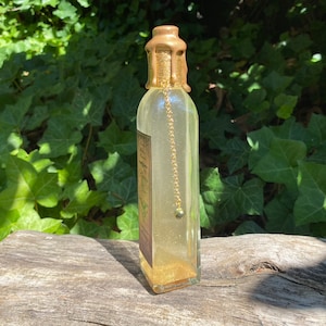 K&ouml;nnte beinhalten: Eine klare Glasflasche mit einem goldfarbenen Wachssiegel und einer Goldkette mit einer kleinen Perle. Die Flasche enth&auml;lt eine goldene Fl&uuml;ssigkeit und Goldglitter. Ein Etikett ist auf der Flasche sichtbar. Die Flasche steht auf einer Holzoberfl&auml;che.