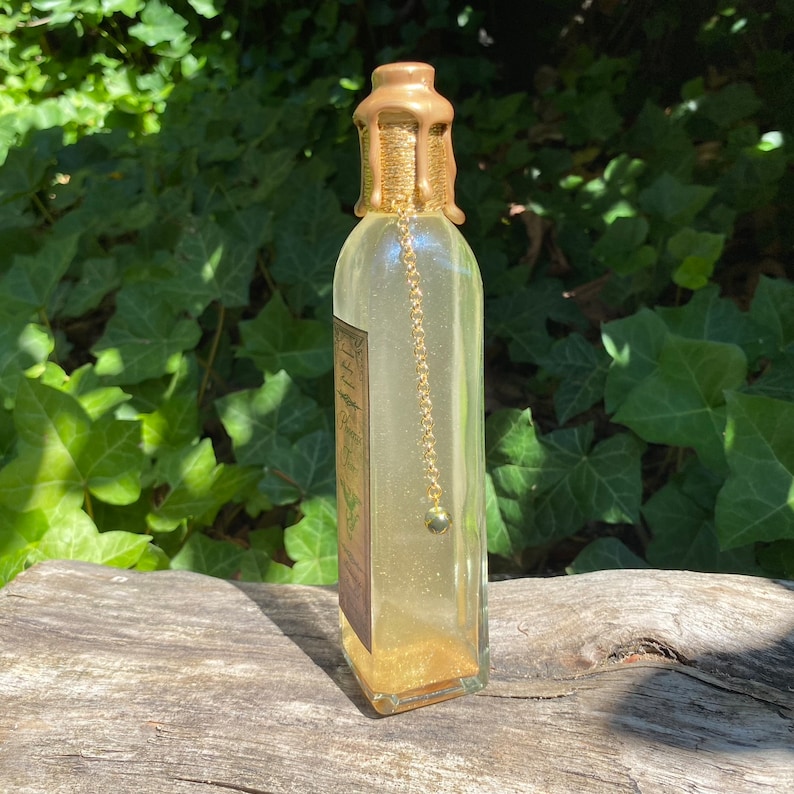 K&ouml;nnte beinhalten: Eine klare Glasflasche mit einem goldfarbenen Wachssiegel und einer Goldkette mit einer kleinen Perle. Die Flasche enth&auml;lt eine goldene Fl&uuml;ssigkeit und Goldglitter. Ein Etikett ist auf der Flasche sichtbar. Die Flasche steht auf einer Holzoberfl&auml;che.